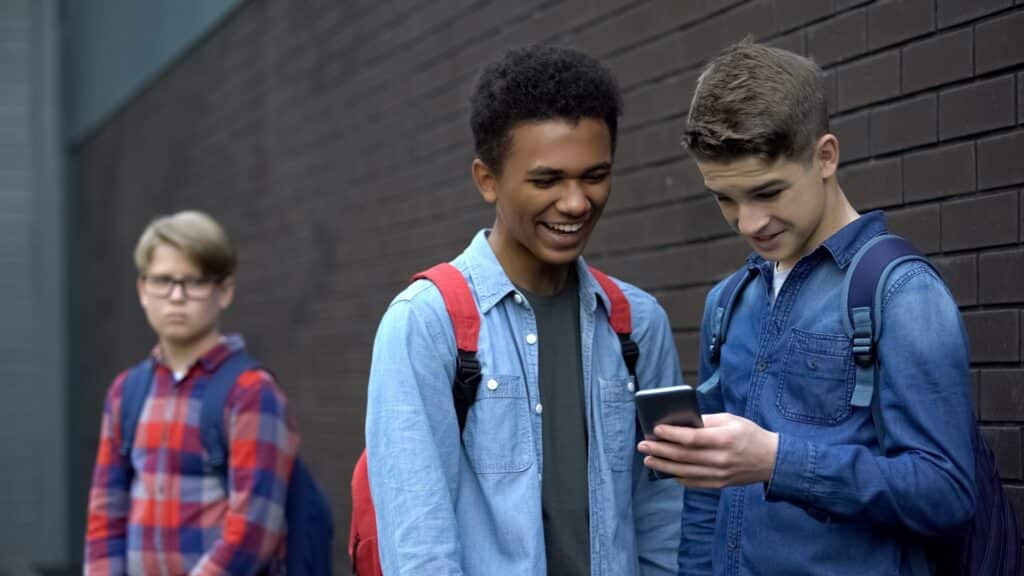 two boys looking amused at a phone with a sad boy in the background