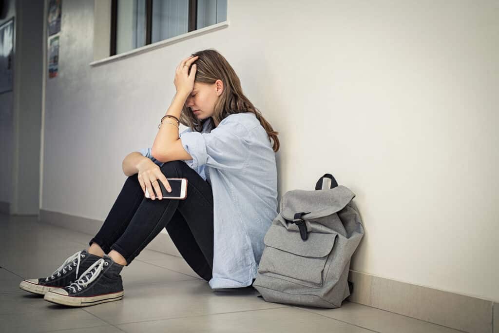 upset teen sitting on floor with phone and hand on her forehead