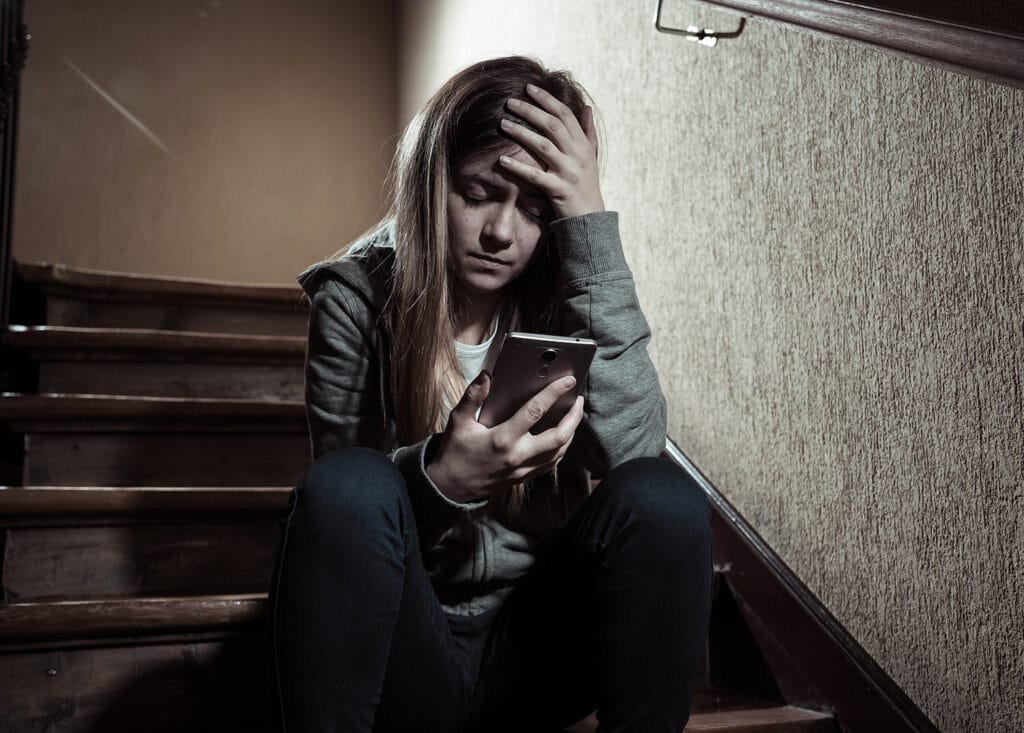 teen girl sitting in dark stairwell looking at phone with hand on her forehead